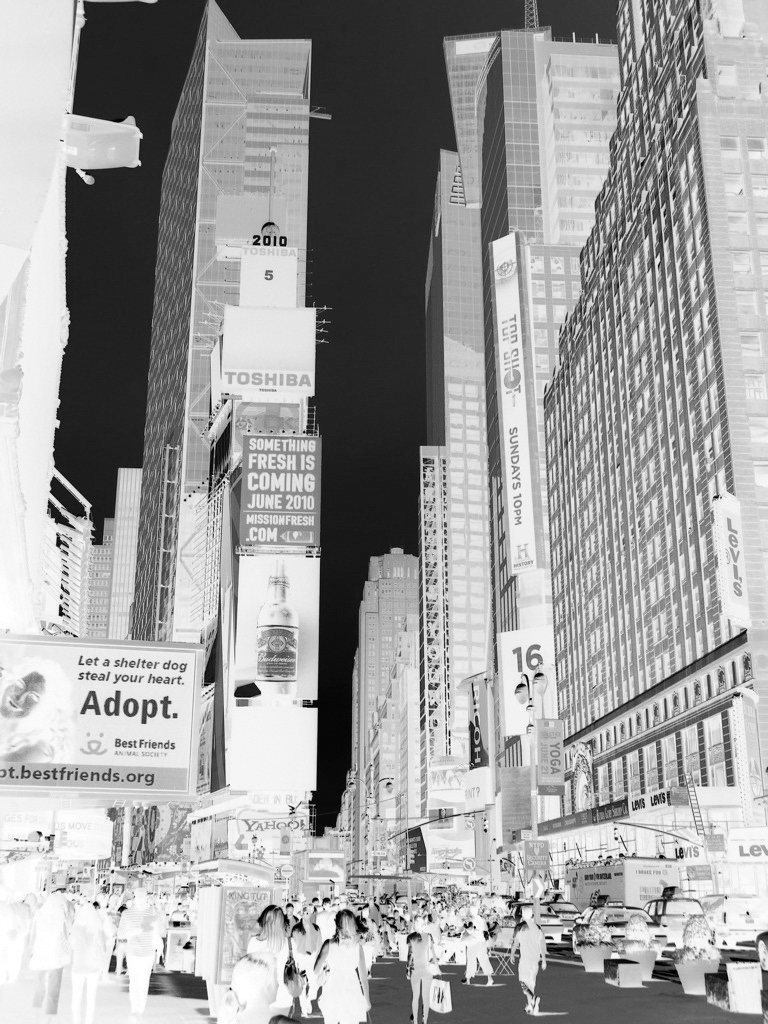 Photographie en noir et blanc d'un carrefour animé de Times Square à New York : taxis, panneaux publicitaires et piétons à un passage clouté.